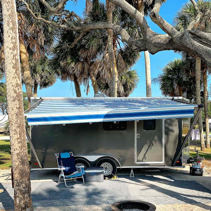 cargo trailer with blue striped awning in campsite with palm trees and sand